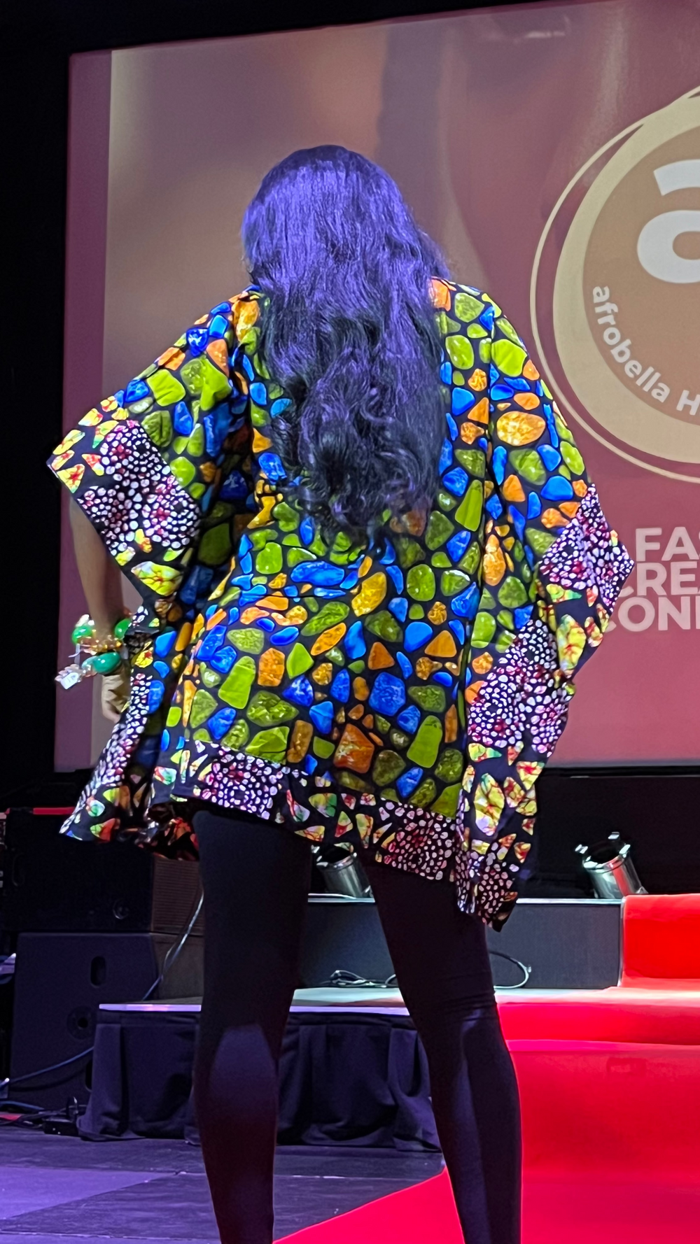 Person wearing a colorful patterned outfit on a stage with 'Afro Bella Hair & Beauty' branding in the background.
